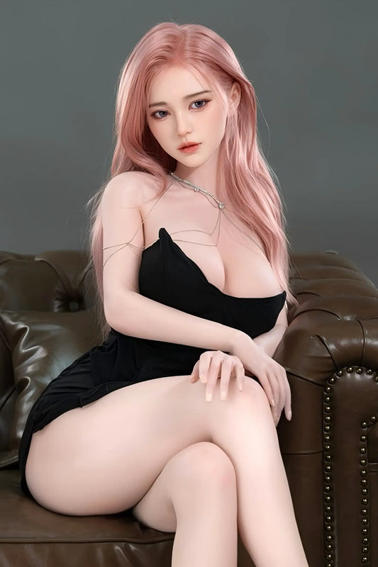 Tyla sex doll bold direct gaze with parted coral lips, deep grey-brown eyes with sculpted brows, long straight rose-pink hair falling over both shoulders, delicate silver body chain across chest, black deep-V strapless mini dress with full round bust prominently displayed and bare thighs fully exposed, slender figure with long legs crossed at knee, one hand resting on thigh, seated on dark brown leather tufted sofa, soft grey studio background – full-body shot – lifelike Asian girl-next-door sex doll by TLD