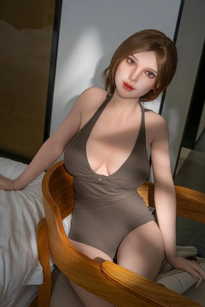 Gadora beautiful sex doll seated in a wooden armchair beside a bed, wearing an olive halter romper