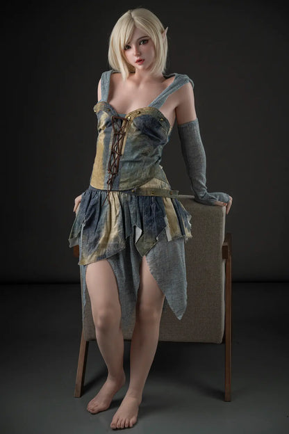 Full-length front view of Elowen blonde elf sex doll standing beside a chair in a lace-up woodland fantasy dress.