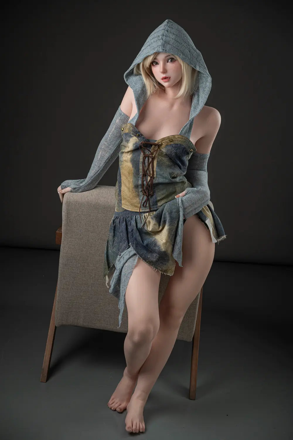 Elowen fantasy cosplay doll standing with crossed ankles beside an armchair, wearing a raised gray-green hood and layered dress.