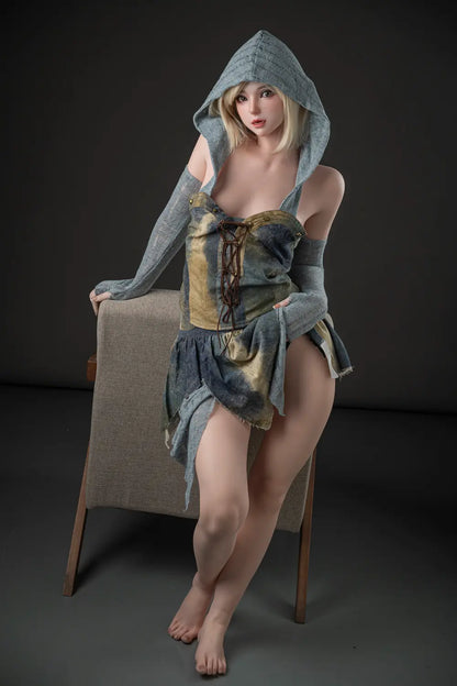 Elowen fantasy cosplay doll standing with crossed ankles beside an armchair, wearing a raised gray-green hood and layered dress.