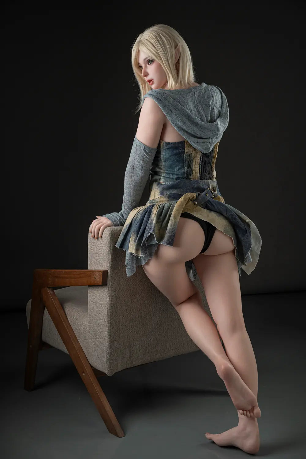 Rear three-quarter view of Elowen fantasy doll leaning on a chair back, looking over her shoulder with her hood resting across her back.