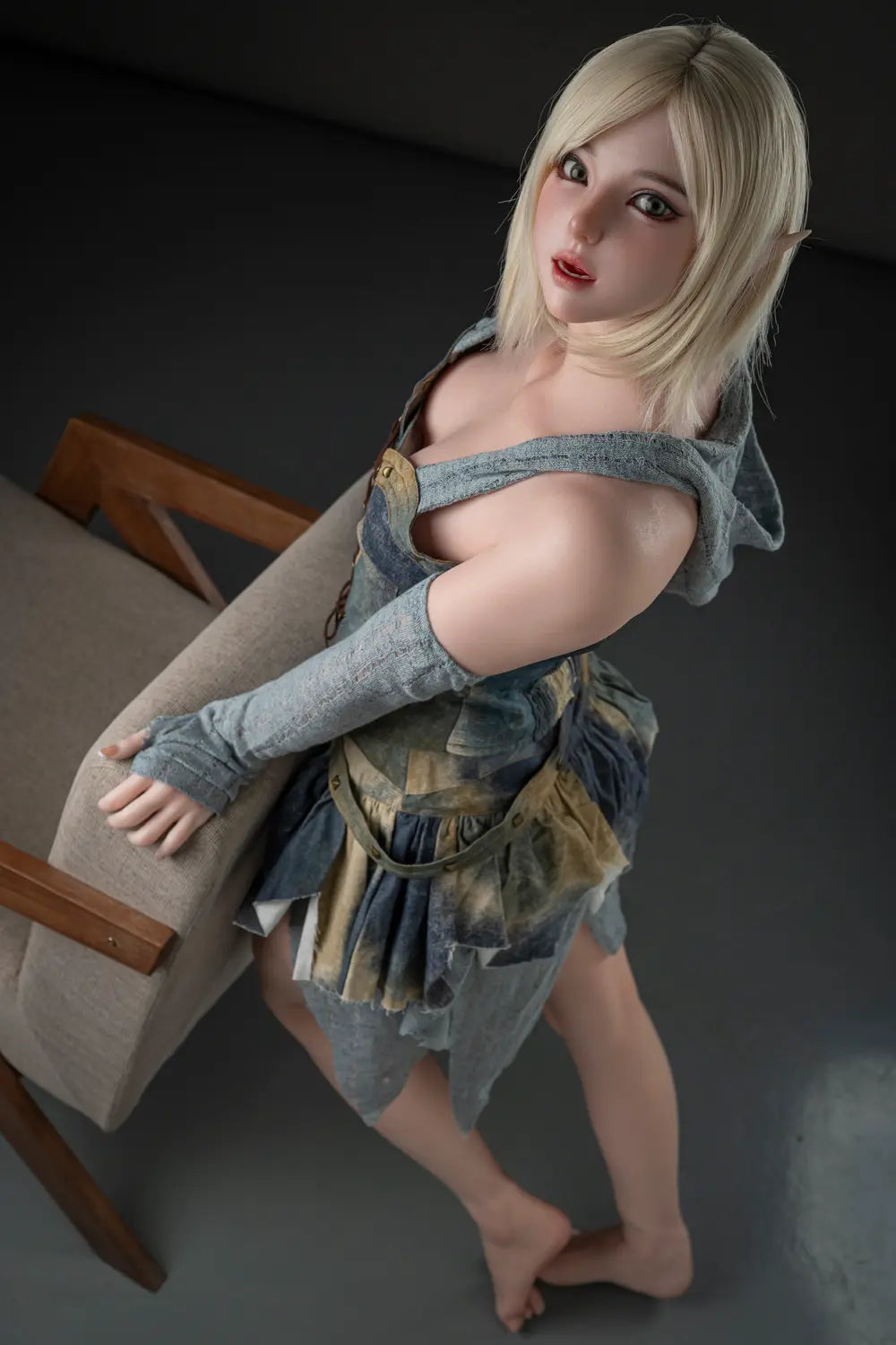 High-angle view of Elowen elf cosplay doll beside an armchair, showing blonde bobbed hair, gray-green sleeves and a layered woodland skirt.