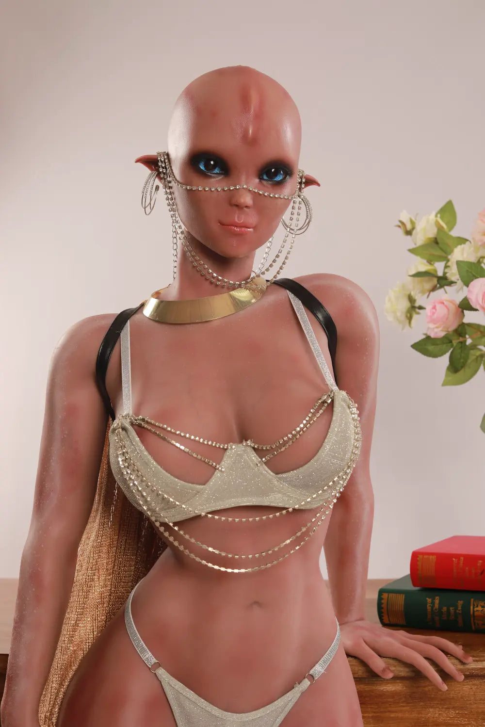 Vaelis pointed-ear alien sex doll in a silver outfit with one hand resting beside stacked books