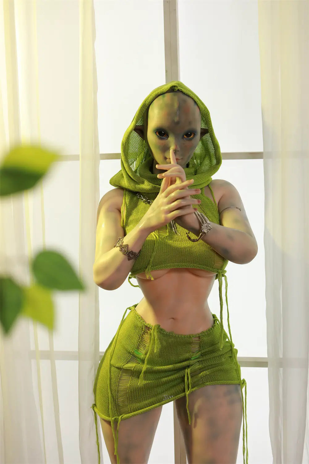 Sylvara woodland sprite sex doll standing in a green hooded outfit with both hands raised beside a bright window