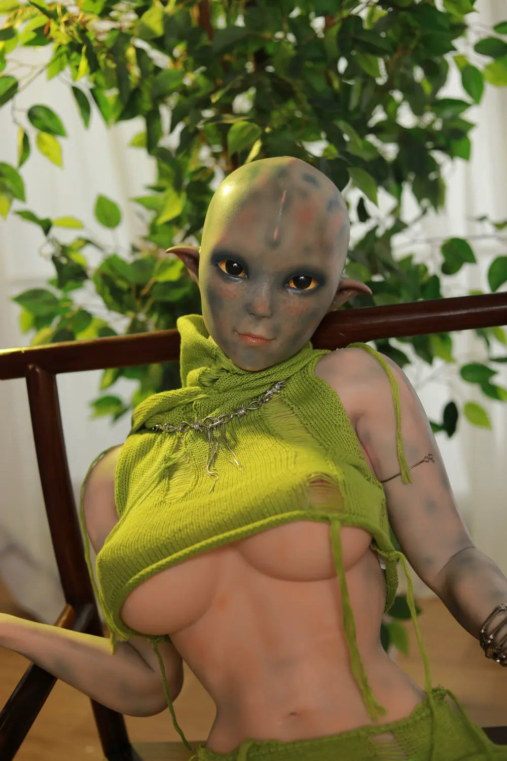 Front seated view of Sylvara sex doll with crossed legs beside leafy plants