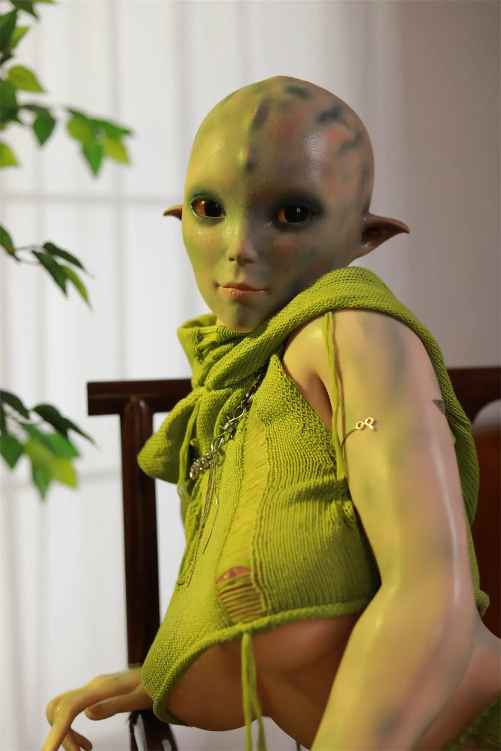 Rear three-quarter view of Sylvara sex doll kneeling on a wooden chair in a green outfit
