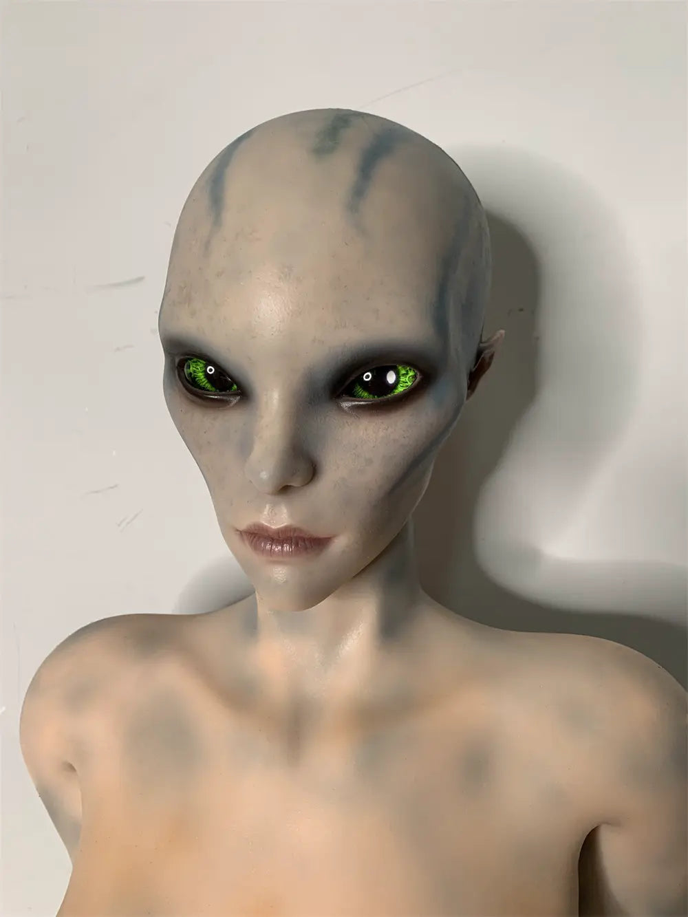 Front factory portrait of Aelyth sex doll showing dark eyes and pale blue markings on the hairless head