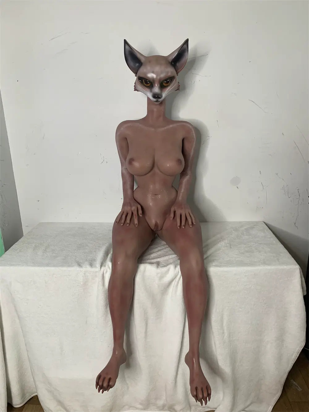 Factory reference photo of unclothed Vesper fox sex doll seated on a white-covered platform